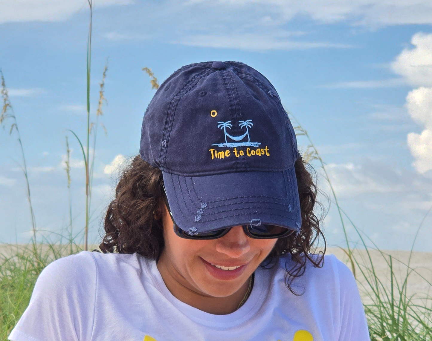 woman sitting on the beach with her head down wearing the distressed dad hat in navy.  Embroidered graphic of the Time to Coast palm tree logo in light blue, yellow time to coast and sun