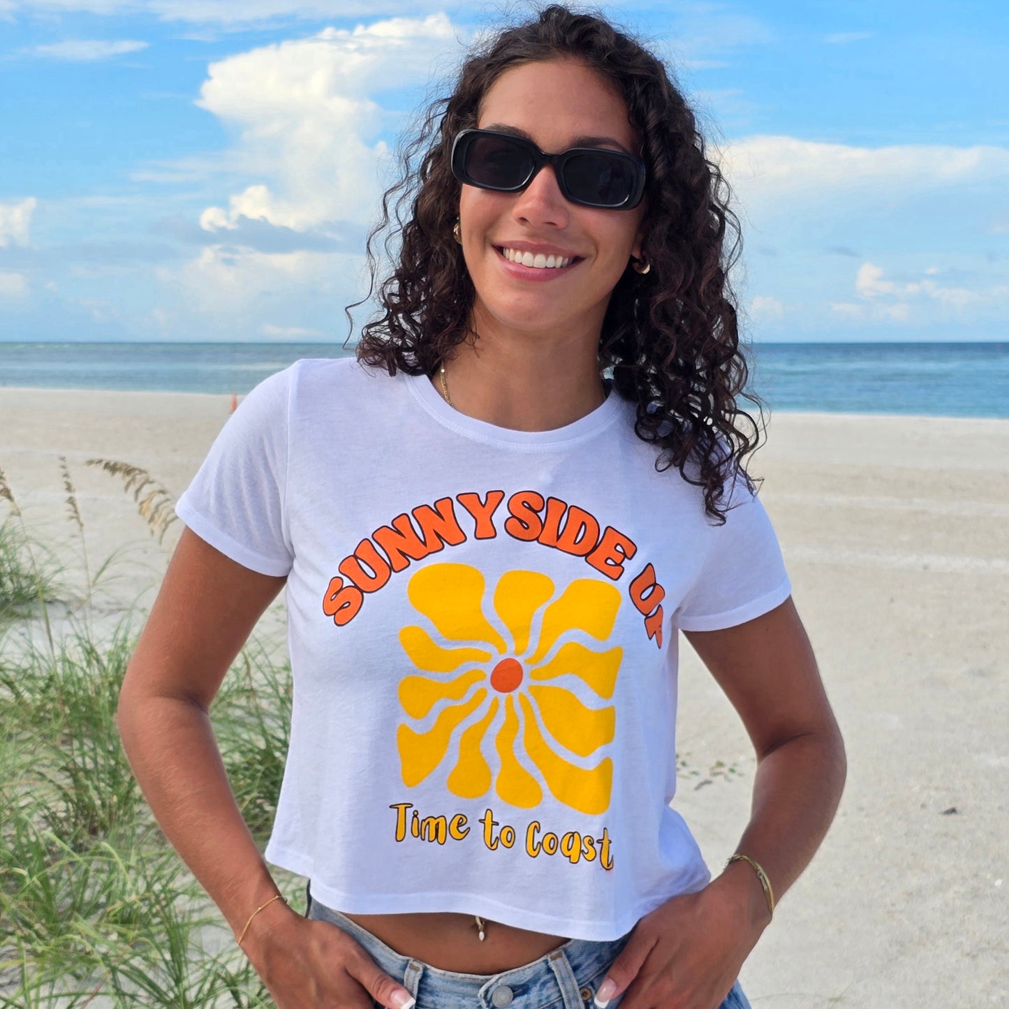 Woman on the beach, wearing sunglasses, denim shorts, and a white Time to Coast Palm Cloth crop tee. Graphic is Sunnyside Up Sun, in yellow and orange colors.  