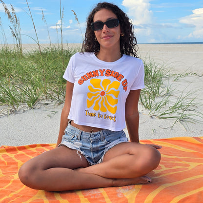 Woman sitting in the sand dunes of the beach, wearing sunglasses, denim shorts, and a white Time to Coast Palm Cloth crop tee. Graphic is Sunnyside Up Sun, in yellow and orange colors.  