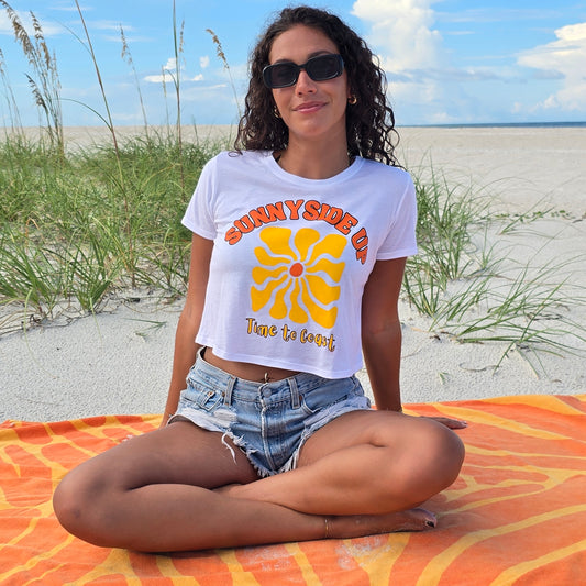 Woman sitting in the sand dunes of the beach, wearing sunglasses, denim shorts, and a white Time to Coast Palm Cloth crop tee. Graphic is Sunnyside Up Sun, in yellow and orange colors.  