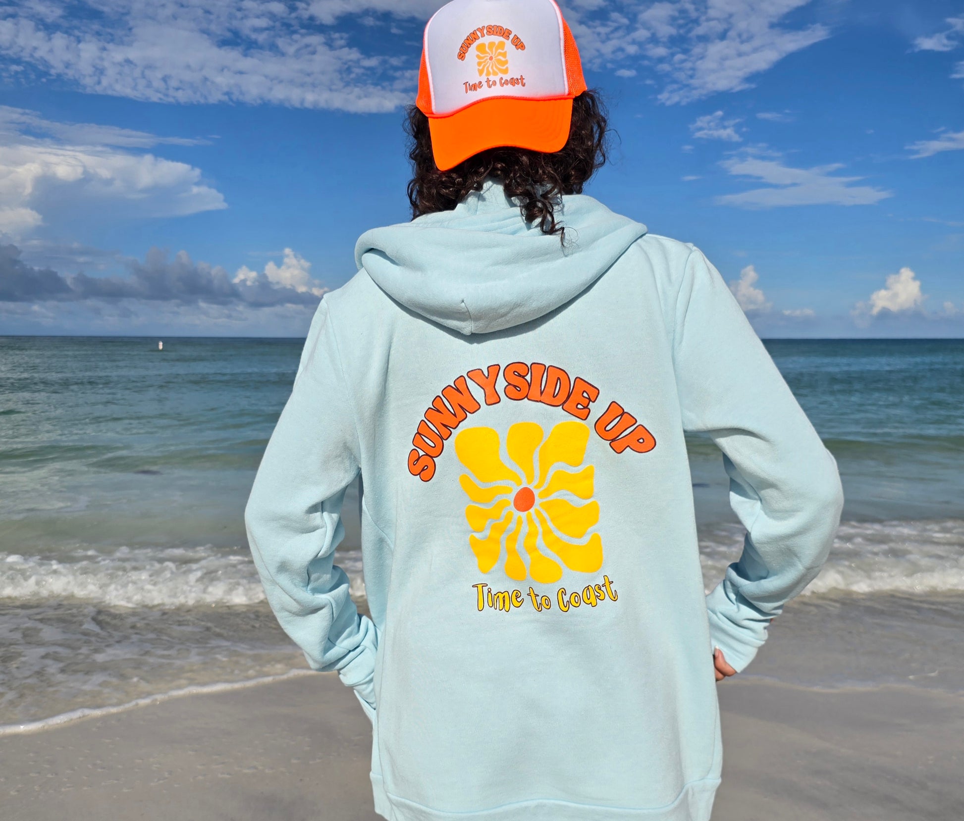 Woman on a beach looking out to the ocean, showing the back of the light blue Florida Fleece Zip Front Hoodie.  The graphic is the Time to Coast Sunnyside Up Sun in orange and yellow. Large graphic on back with small left chest graphic.  One sleeve has the words where summer never ends