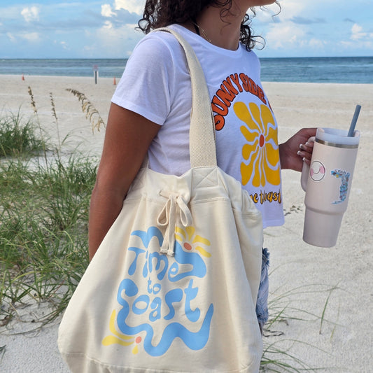 Woman on the beach wearing Sunglasses, the Palm Cloth sunny side up crop tee, denim shorts and the hobo bag on her shoulder, shows size of  bag. The Florida Fleece Hobo Beach Bag, with drawstring closure. Creme colored with Time to Coast Hippy Wave Graphic in light blue and yellow. 