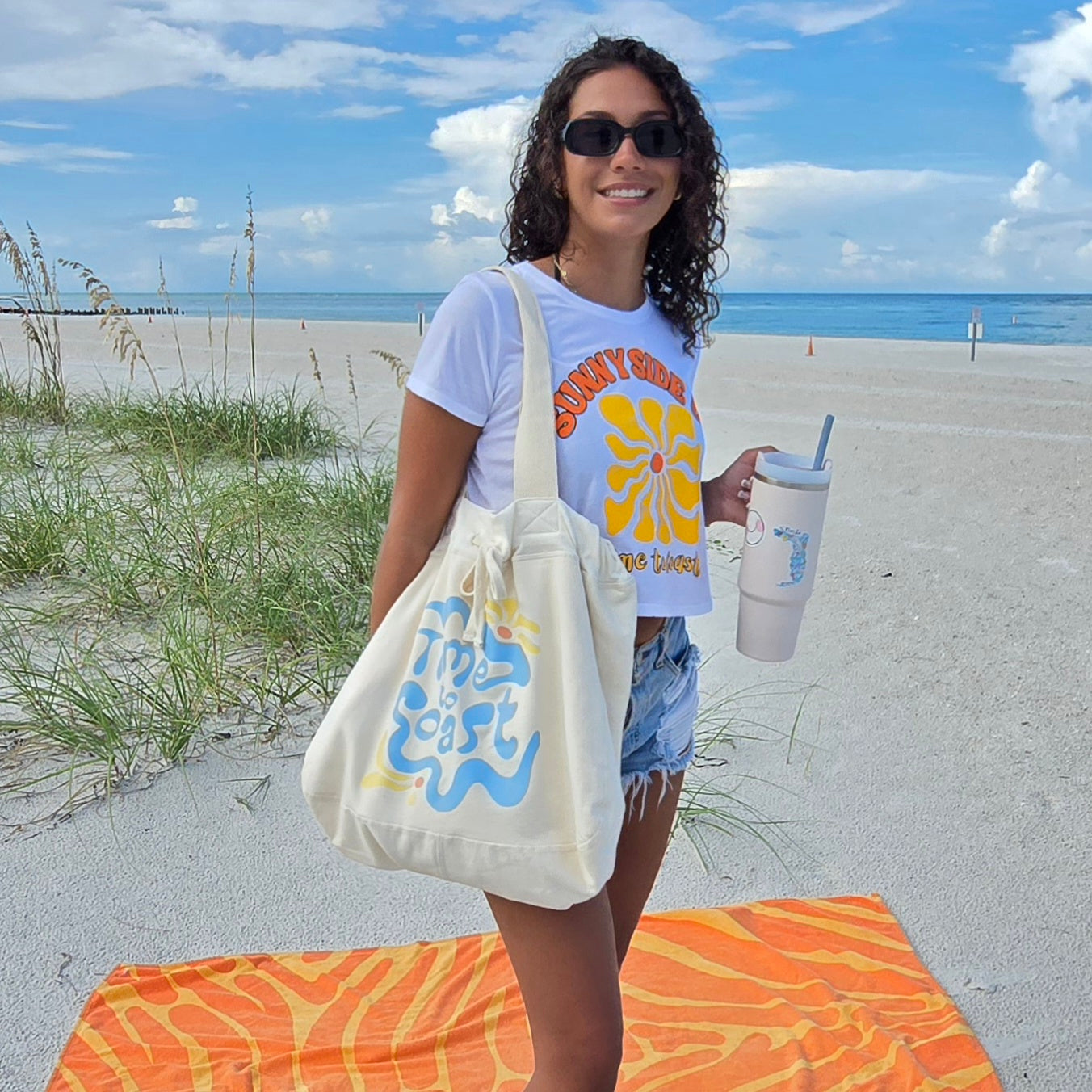 Woman on the beach wearing Sunglasses, the Palm Cloth sunny side up crop tee, denim shorts and the hobo bag on her shoulder, shows size of  bag. The Florida Fleece Hobo Beach Bag, with drawstring closure. Creme colored with Time to Coast Hippy Wave Graphic in light blue and yellow. 