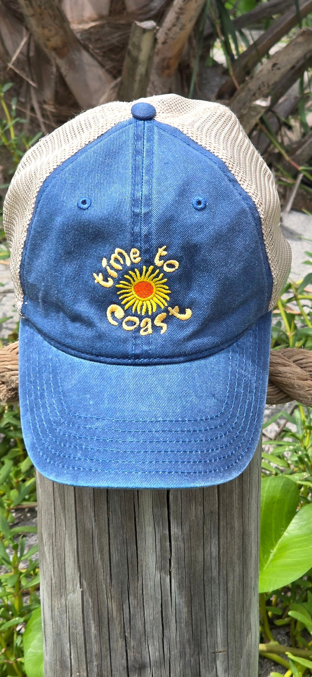 Hat on a beach pole of the Time to sun low profile trucker twill hat with tan mesh back in washed navy. Graphic is sun with time to coast circling the sun. Embroidery is yellow.