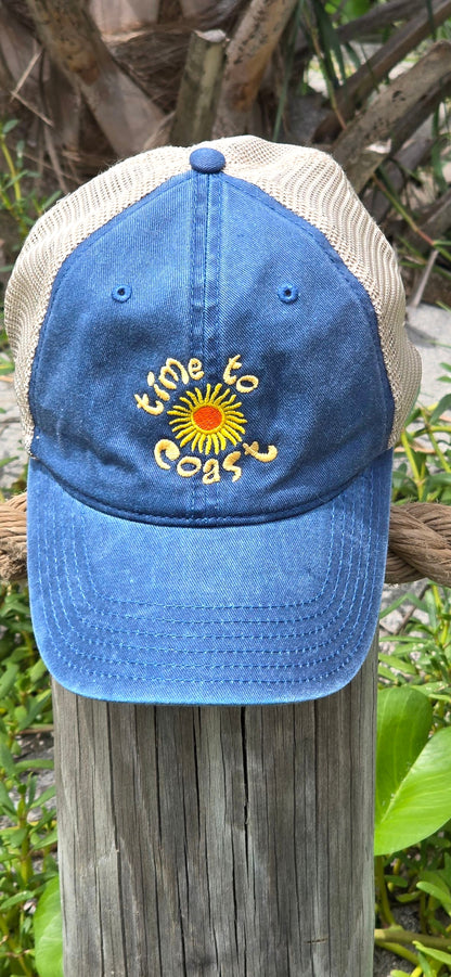 Hat on a beach pole of the Time to sun low profile trucker twill hat with tan mesh back in washed navy. Graphic is sun with time to coast circling the sun. Embroidery is yellow.