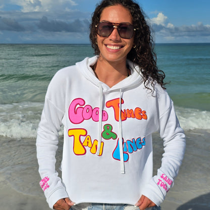 Woman standing on the beach by the ocean wearing sunglasses, denim shorts and a Florida Fleece White Cropped Hoodie Graphic Top.  Graphic is the Time to Coast Good Times & Tan Lines Slogan.  Graphic is in multi colorful words.  Sleeves have hot pink words of sandy toes & sun kissed nose. 