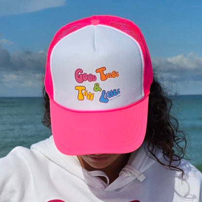 Woman on a beach wearing a foam high profile trucker hat.  Graphic is printed with Time to Coast Good Times and Tan Lines slogan. Hat color is bright pink bill and mesh back with white front. 