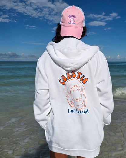 Woman on a beach looking out to the ocean, showing the back of the White Florida Fleece Zip Front Hoodie.  The Coastie Manatee graphic, white manatee and orange. Large graphic on back with small left chest graphic.  One sleeve has the words where summer never ends.