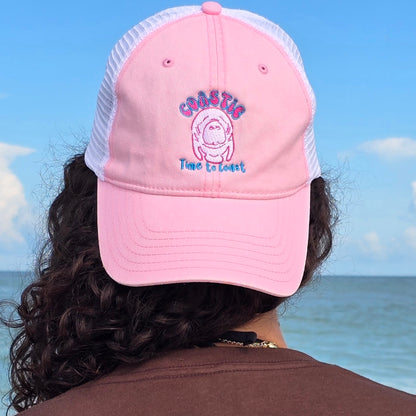 Woman on the beach looking out at the ocean, wearing backwards the Coastie Manatee embroidered twill low profile trucker hat. Color of hat is washed pink twill with a white embroidered manatee and tan mesh back.