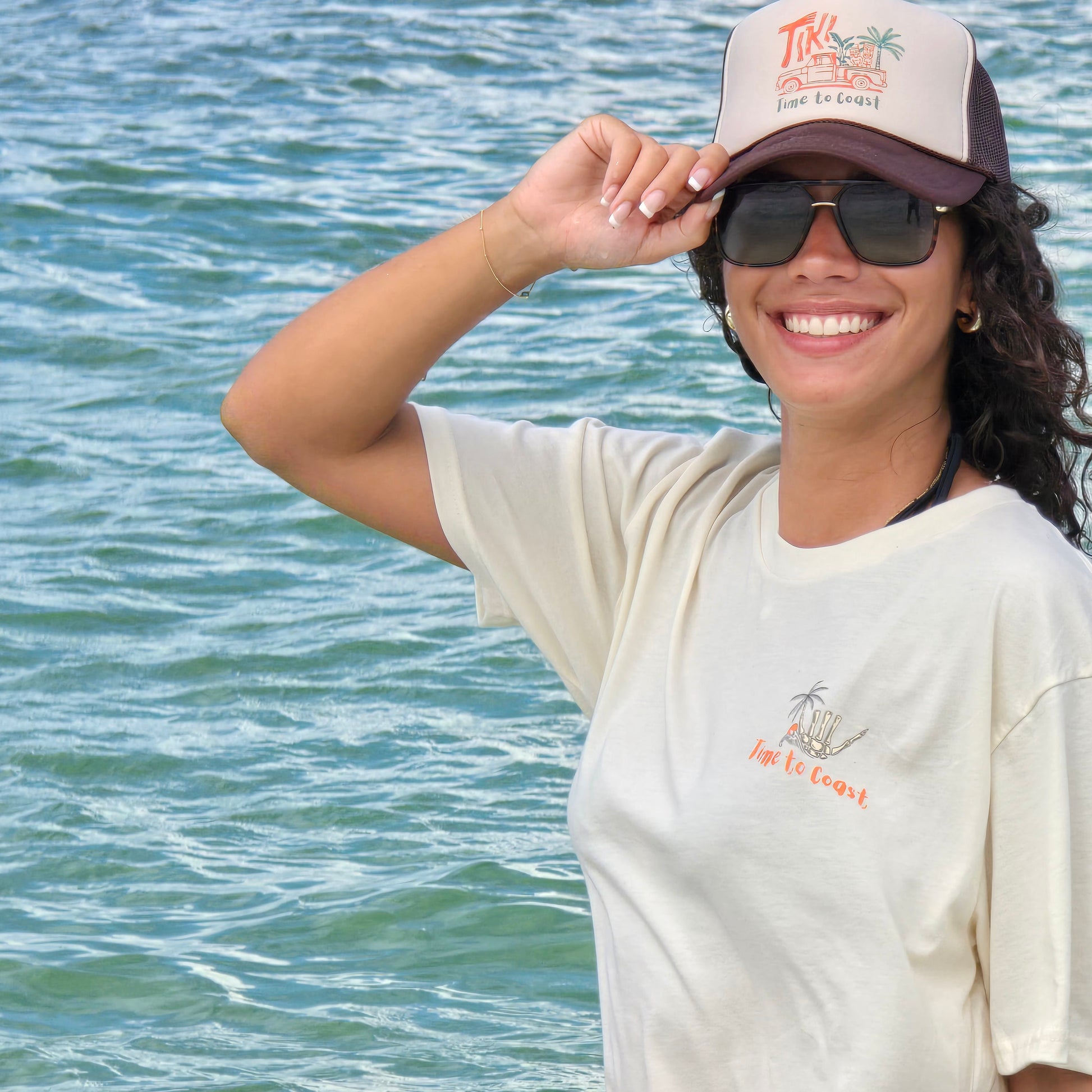 Woman in ocean wearing a foam high  profile trucker hat.  Graphic is printed with Time to Coast Tiki Truck. Hat color is beige and brown with a brown mesh back.