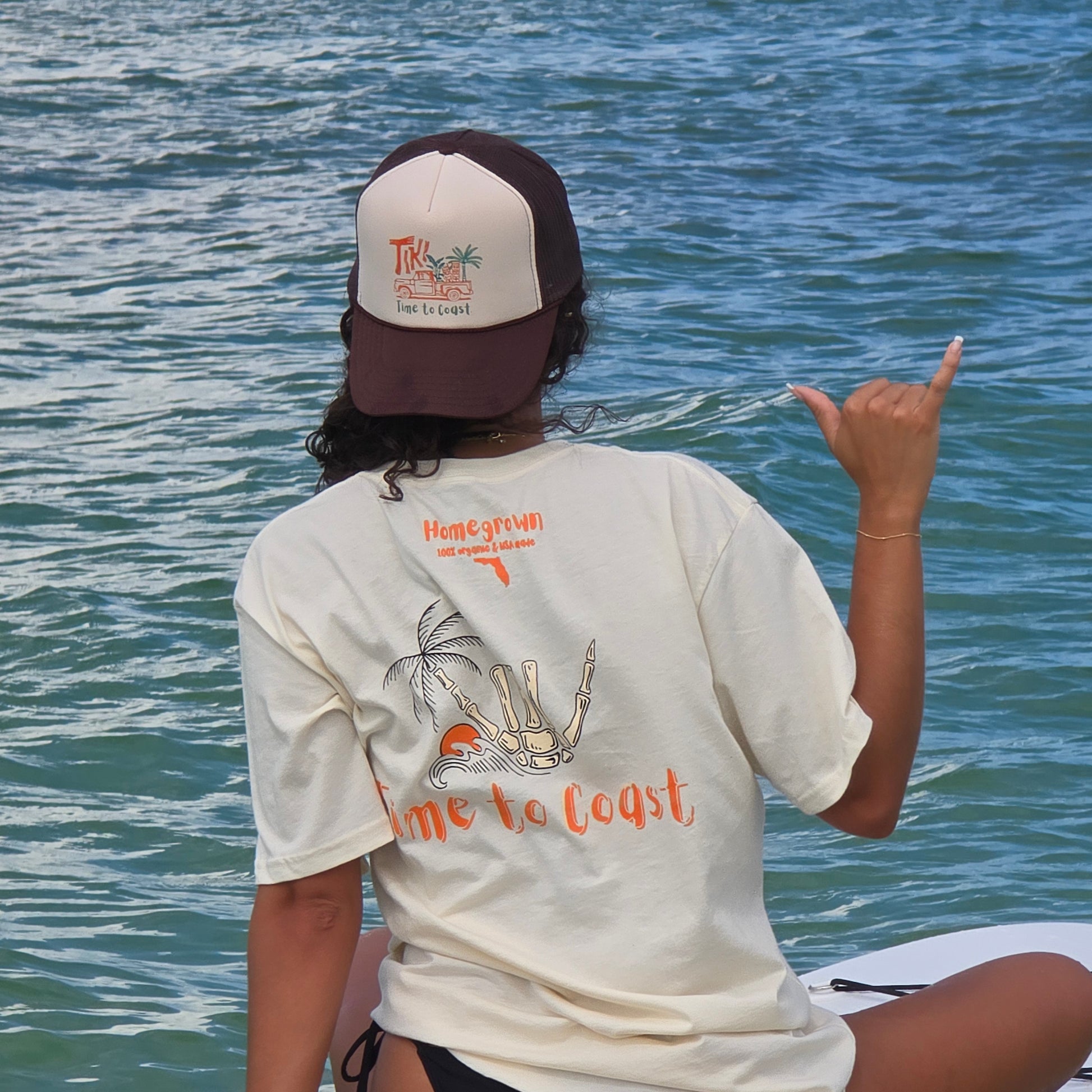 Woman on a sup board, in the ocean, wearing a foam high profile trucker hat backwards.  Graphic is printed with Time to Coast Tiki Truck. Hat color is beige and brown with a brown mesh back.