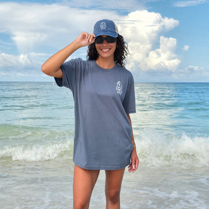 woman standing on the beach ocean shore wearing sunglasses and and a washed navy trucker hat. She is wearing a gray blue unisex  graphic  t-shirt in organic cotton made in USA. Homegrown t-shirt showing the small left chest graphic of swell surf waves.  Also has large back graphic of surf waves and time to coast logo.